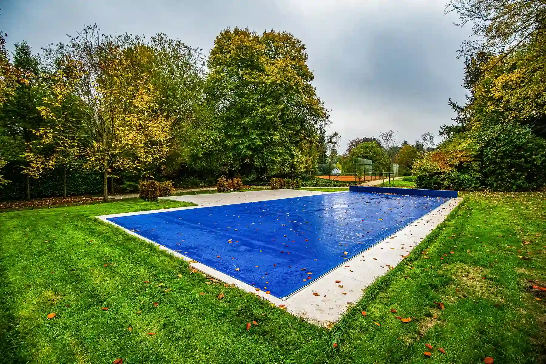 Chez-Elizabeth-et-Jacques-3 Piscine couverte d'une bâche bleue entourée de pelouse verte et d'arbres aux feuilles jaunissantes.