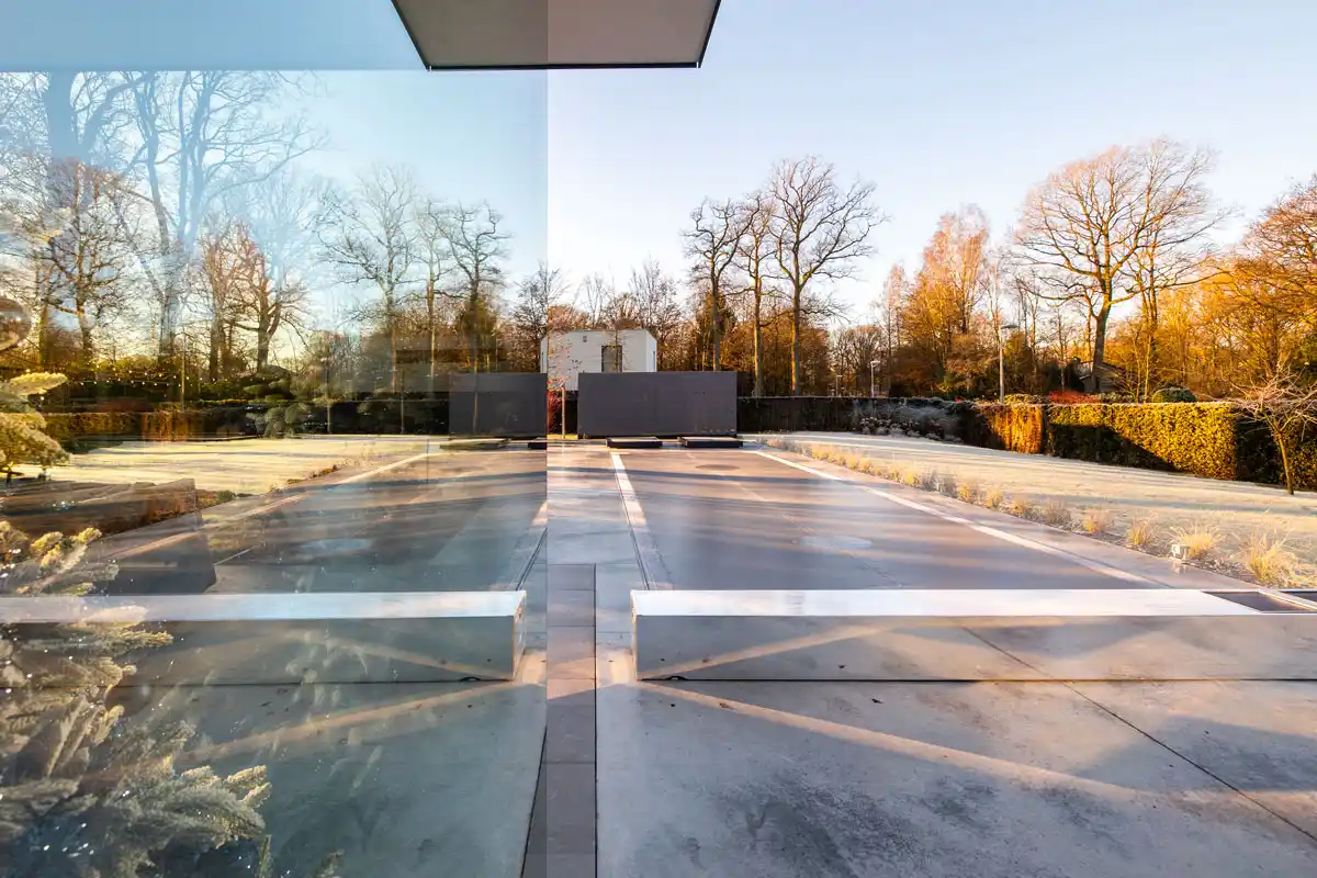 alice-et-paul-gallery-01 Allée en béton avec des reflets de verre, bordée de haies et d'arbres nus sous un ciel clair et ensoleillé.