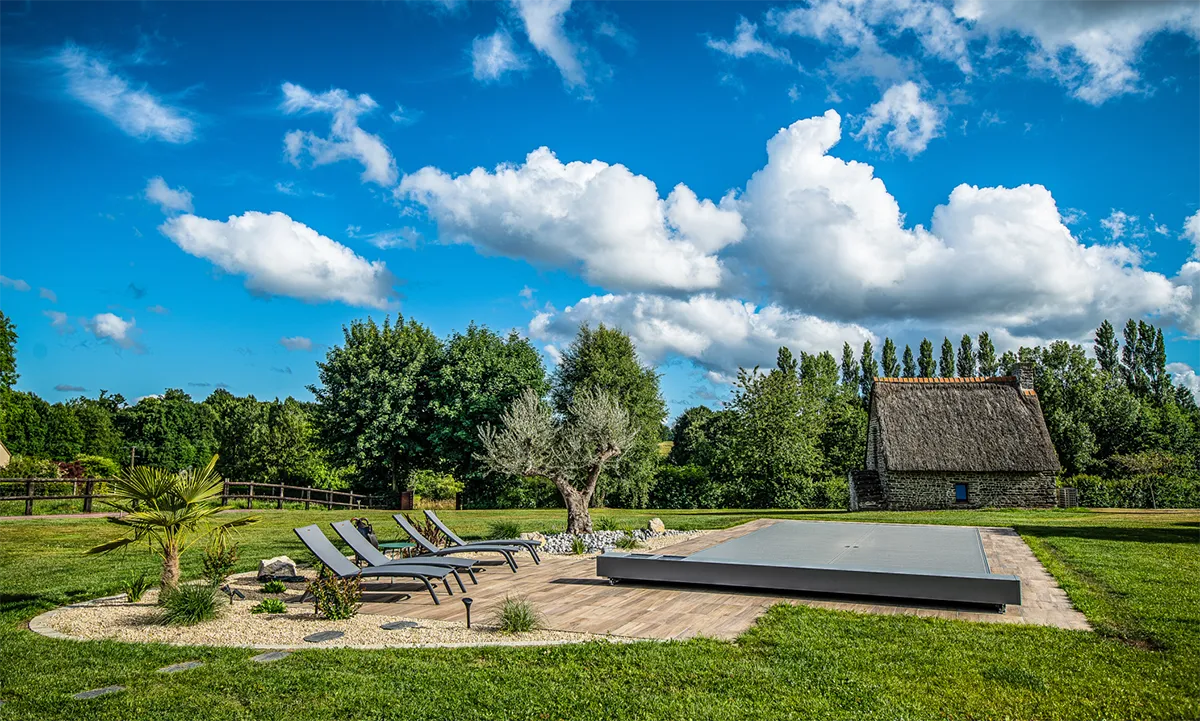 Des chaises longues noires entourent une piscine couverte dans un jardin verdoyant, avec un arbre et une maison en pierre en arrière-plan.