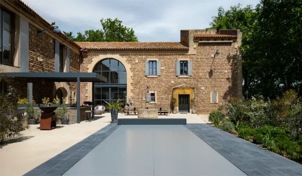 modeles-automatique-3 Cour intérieure avec piscine couverte, entourée de bâtiments en pierre, mobilier de jardin moderne et végétation luxuriante sous ciel nuageux.