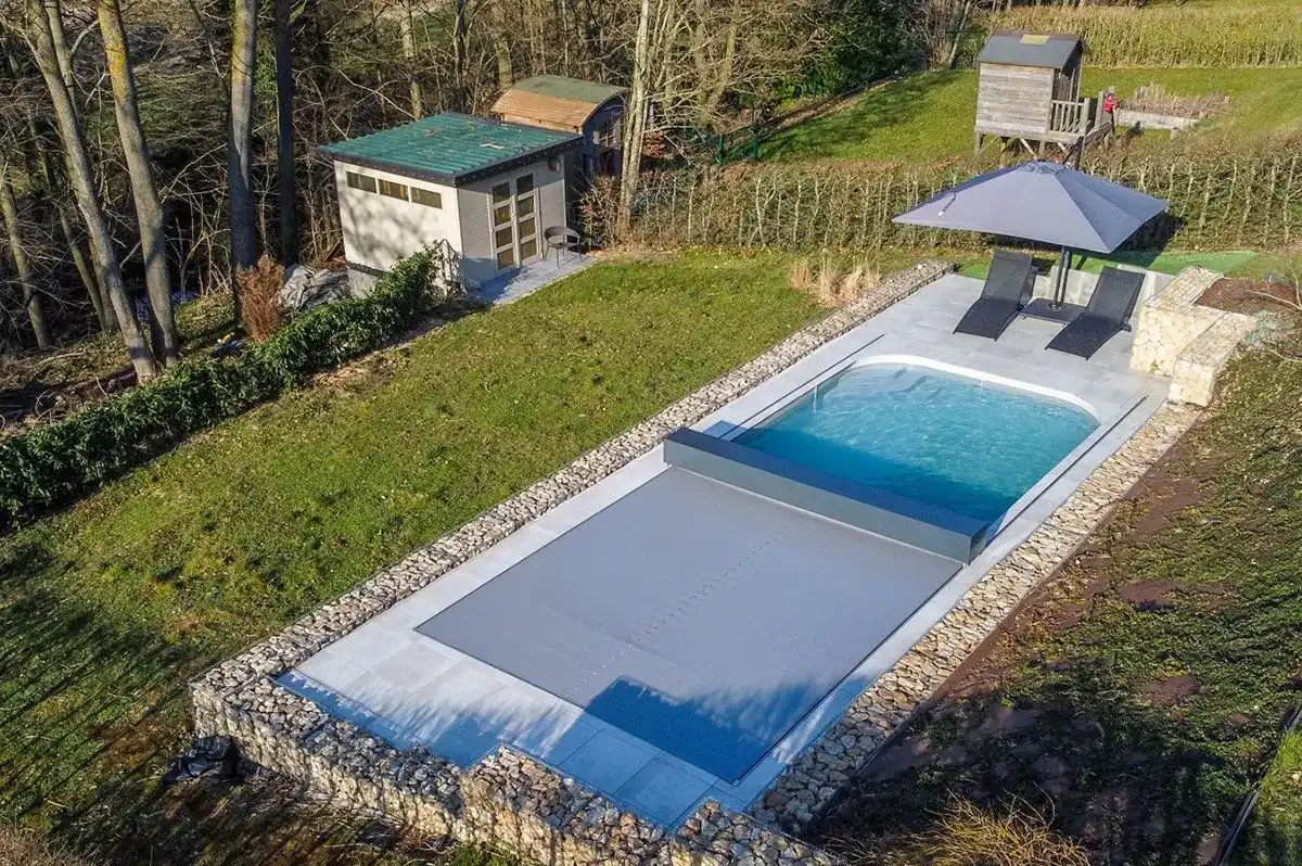 Piscine extérieure entourée de pierres avec couverture grise, deux chaises longues noires sous un parasol, et cabanon en arrière-plan.