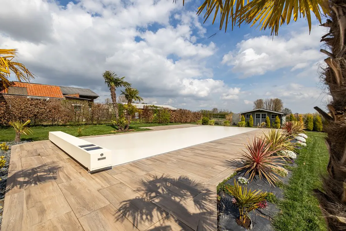Une terrasse en bois entoure une piscine couverte, avec des palmiers et des buissons colorés autour, sous un ciel partiellement nuageux.