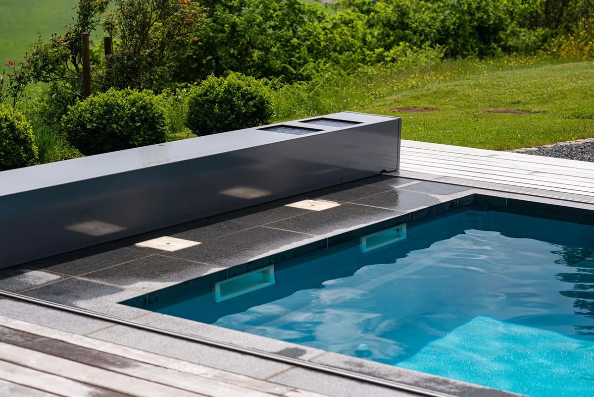 Piscine extérieure avec margelles en pierre grise, entourée de verdure et d'une plateforme en bois clair.