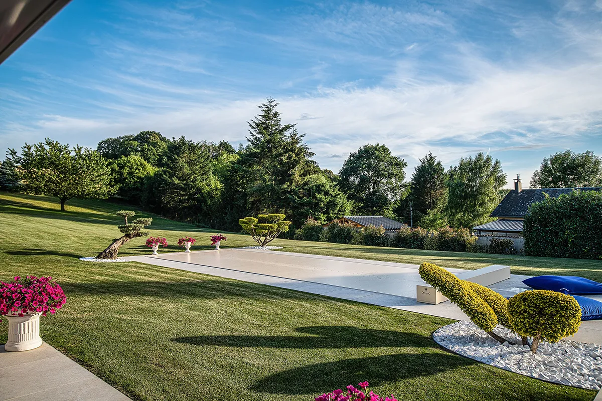 Jardin avec pelouse verte, piscine couverte, arbustes taillés et pots de fleurs roses sous un ciel bleu.