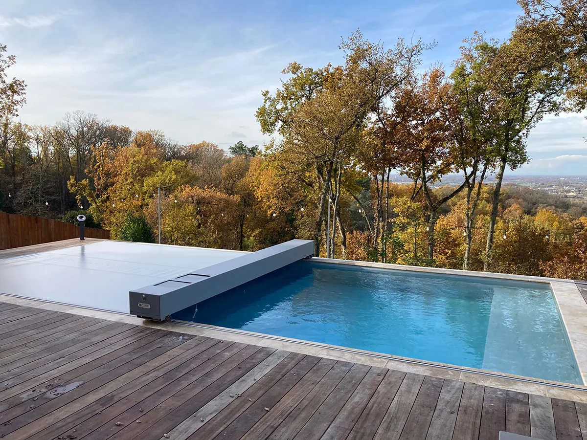 Piscine avec couverture grise sur terrasse en bois, entourée d'arbres aux feuilles automnales et ciel partiellement nuageux.