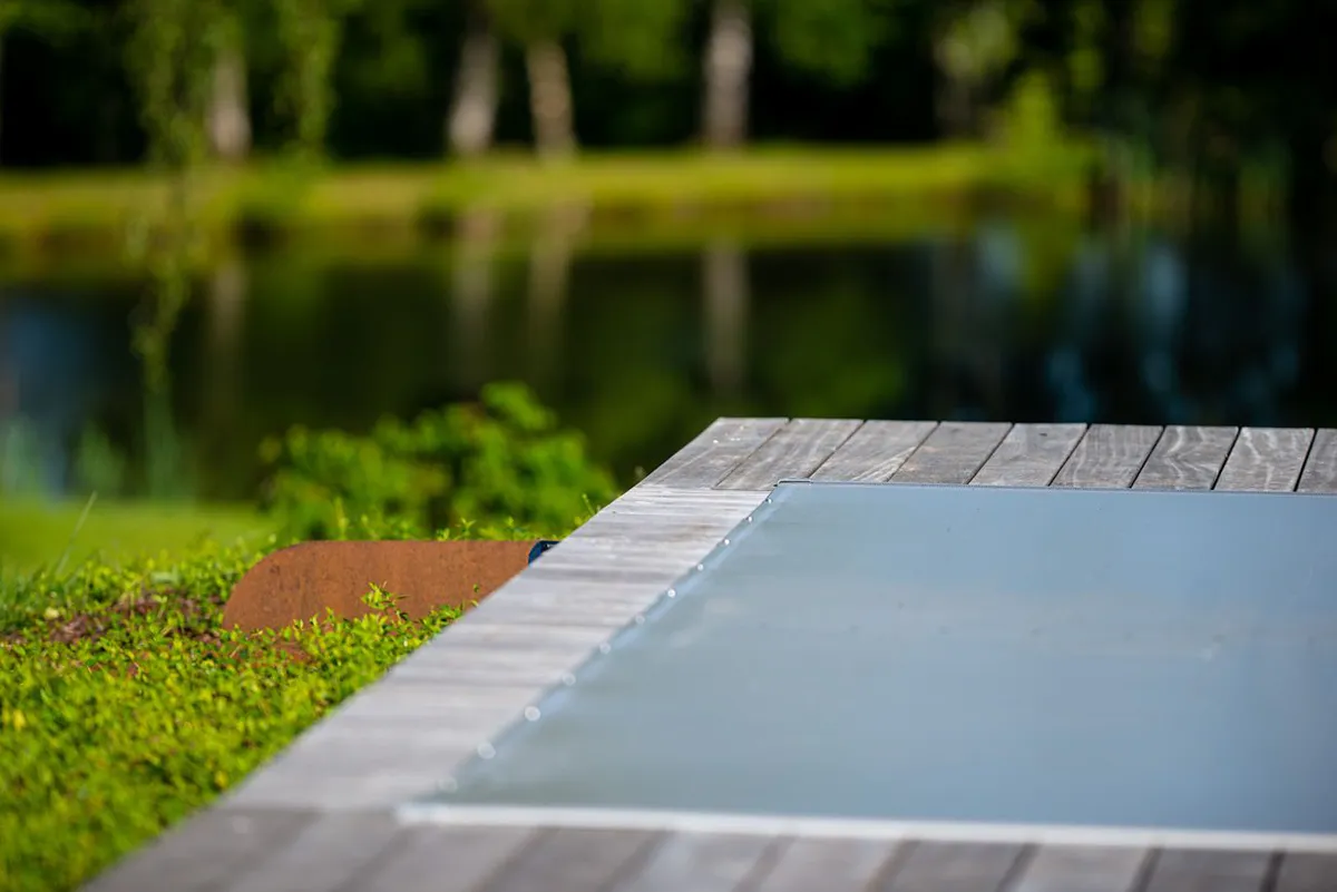 Bord de piscine en bois avec couverture grise, entouré de verdure et d'un étang en arrière-plan.