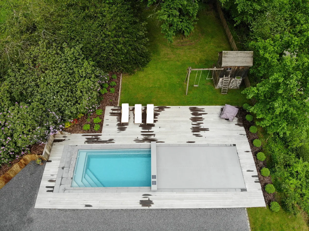 Vue aérienne d'une piscine rectangulaire avec couverture grise sur terrasse en bois clair, entourée de verdure et d'un espace de jeux pour enfants.