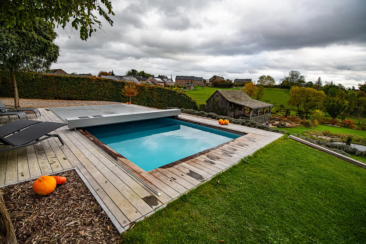 Piscine extérieure avec couverture grise entourée de terrasse en bois, citrouilles et chaises longues, paysage de campagne en arrière-plan.