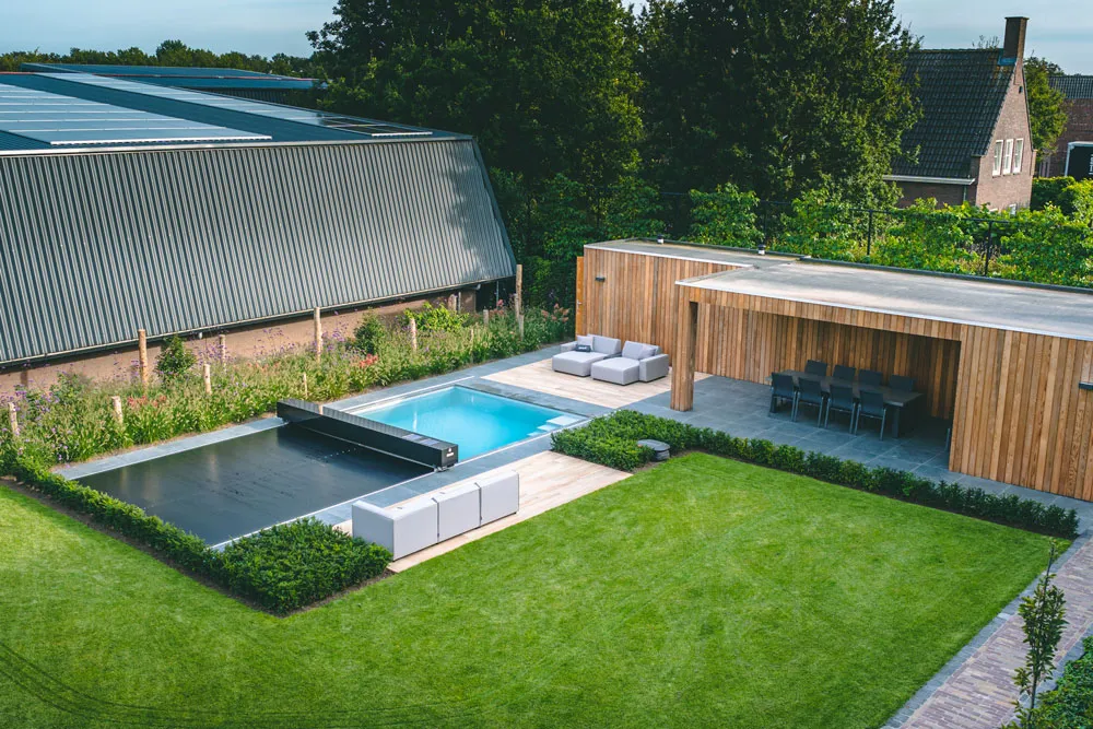 Piscine rectangulaire avec couverture noire, entourée de pelouse verte, terrasse en bois avec salon extérieur et table à manger, bâtiment à toit métallique.