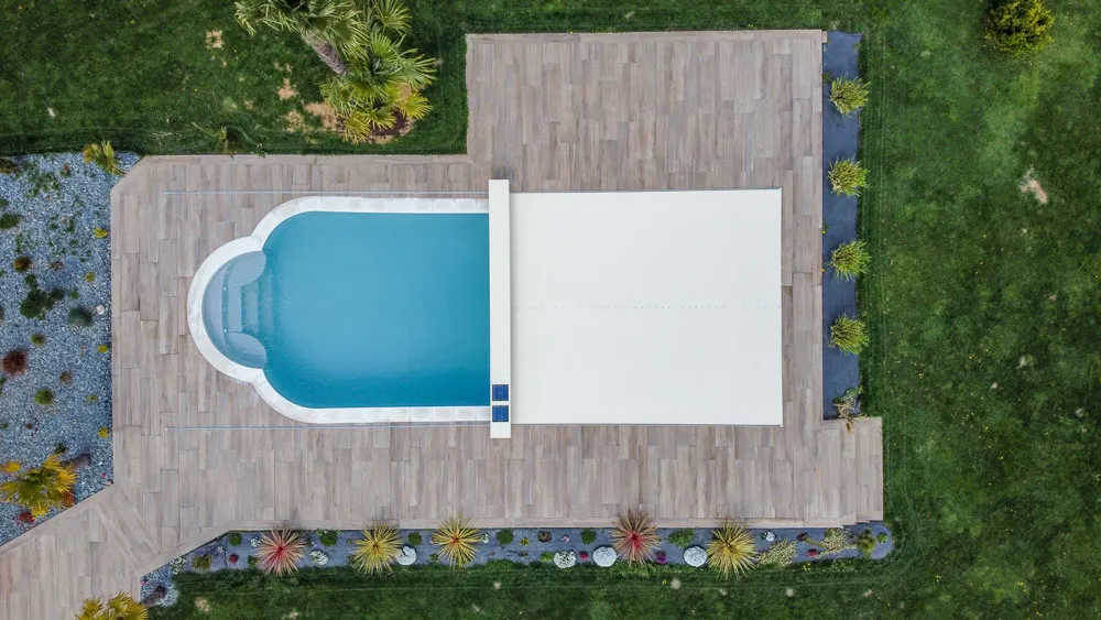 Vue aérienne d'une piscine bleue en forme de L partiellement couverte, entourée de terrasses en bois et de pelouses vertes.