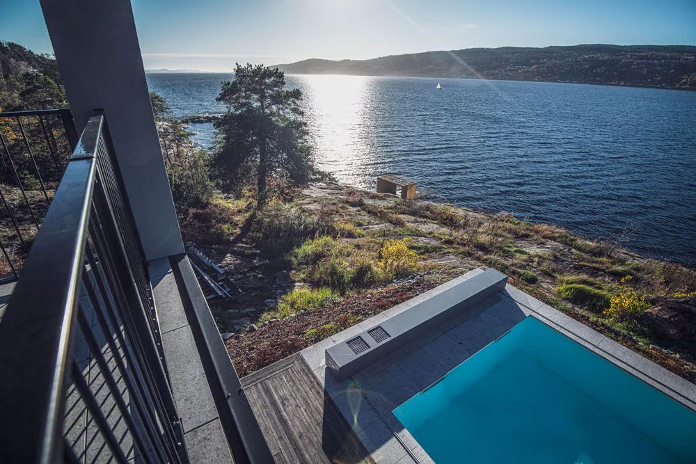 Vue d'une piscine avec terrasse en bois, bordée par un paysage naturel et un lac en arrière-plan, éclairé par le soleil.