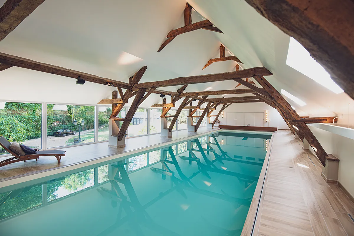 Piscine intérieure avec eau turquoise, entourée de poutres en bois et grandes fenêtres donnant sur un jardin verdoyant.