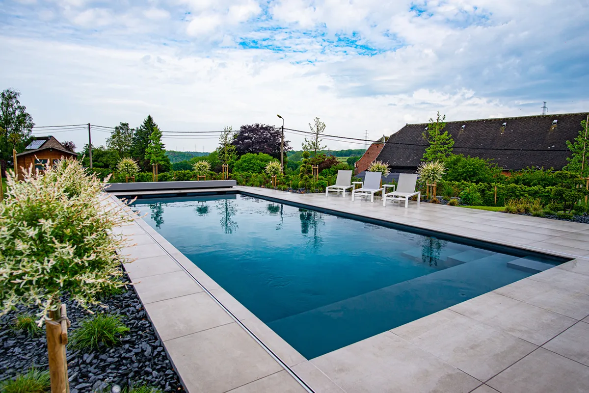 Piscine rectangulaire entourée de dalles grises et de chaises longues blanches, avec des arbres et un ciel partiellement nuageux en arrière-plan.