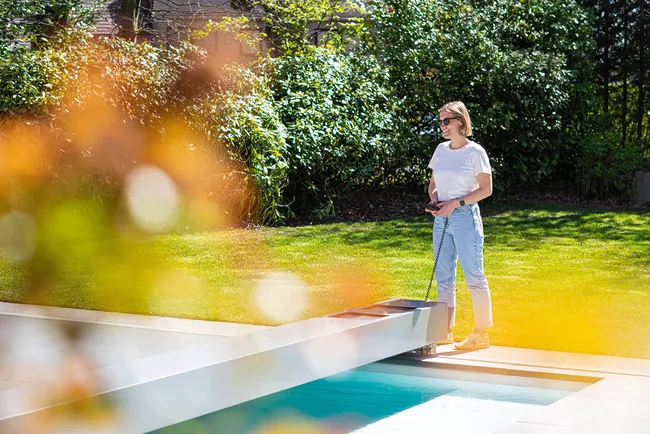 Une personne en t-shirt blanc et jeans se tient près d'une piscine, entourée de verdure et de buissons.