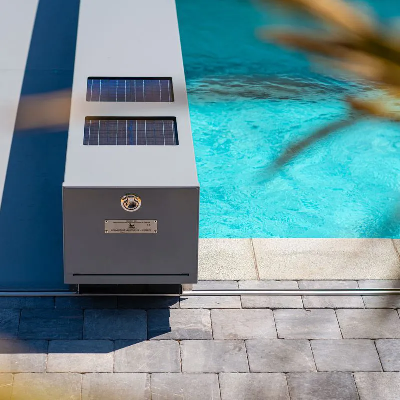 Couvercle de piscine gris avec panneaux solaires intégrés, adjacent à une piscine d'eau bleue claire et un sol en dalles grises.