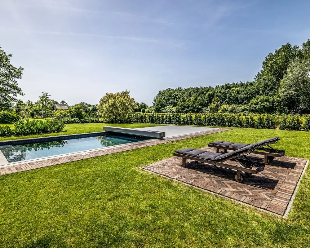 Piscine rectangulaire avec couverture grise entourée de pelouse verte, deux transats noirs sur une terrasse en bois, arbres et haies en arrière-plan.