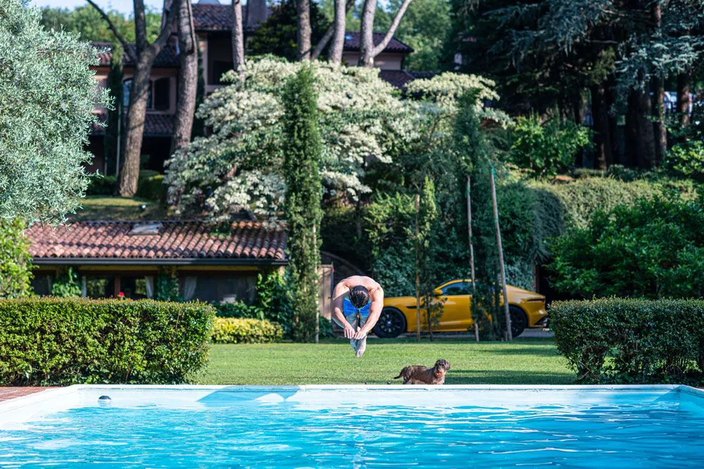 Un homme plonge dans une piscine bleue entourée de haies vertes, avec un chien et une voiture jaune en arrière-plan.