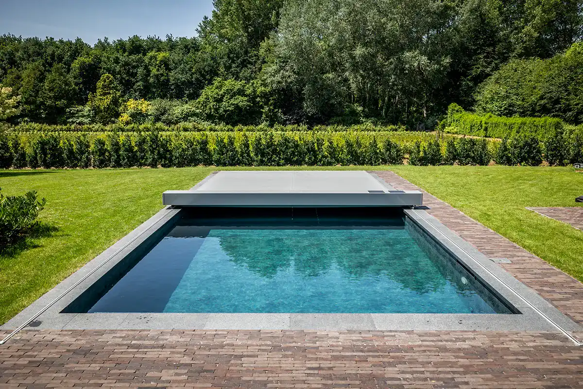 Piscine rectangulaire avec couverture grise, entourée de pelouse verte et de haies, ciel dégagé en arrière-plan.