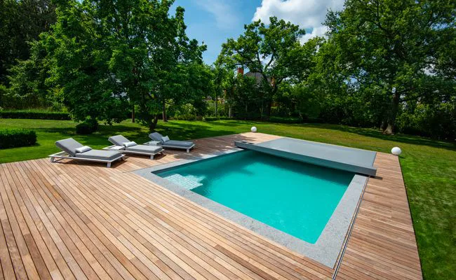 Piscine rectangulaire entourée de terrasse en bois avec quatre transats gris, située dans un jardin verdoyant avec arbres.
