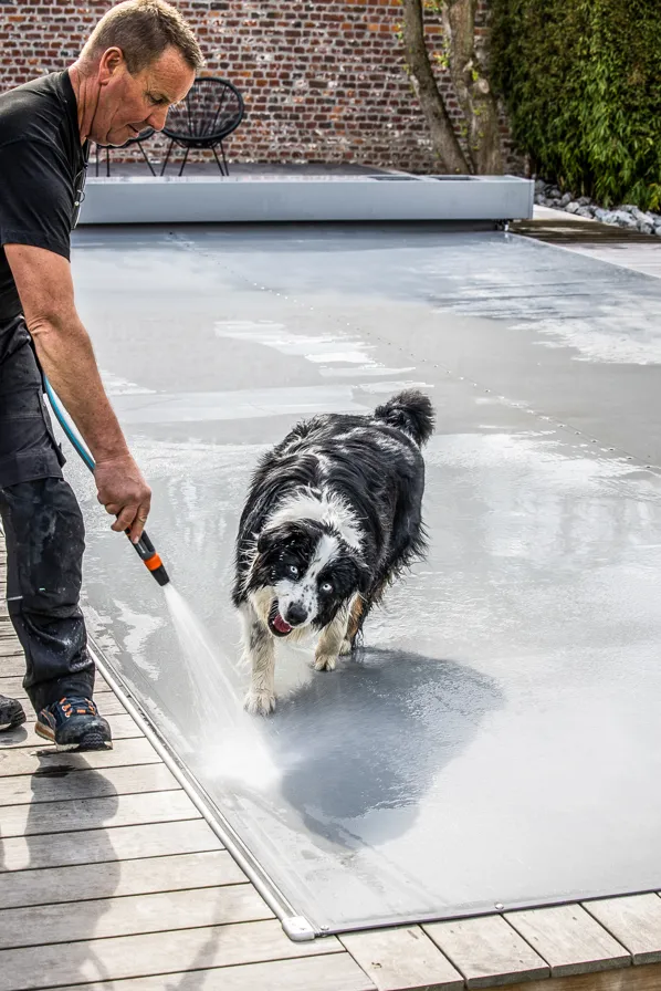 Un homme nettoie une couverture de piscine grise avec un tuyau d'arrosage, accompagné d'un chien noir et blanc.