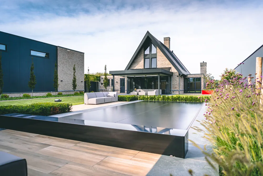 Maison moderne avec toit en pente, terrasse en bois, piscine couverte, canapé blanc et jardin fleuri sous un ciel bleu.