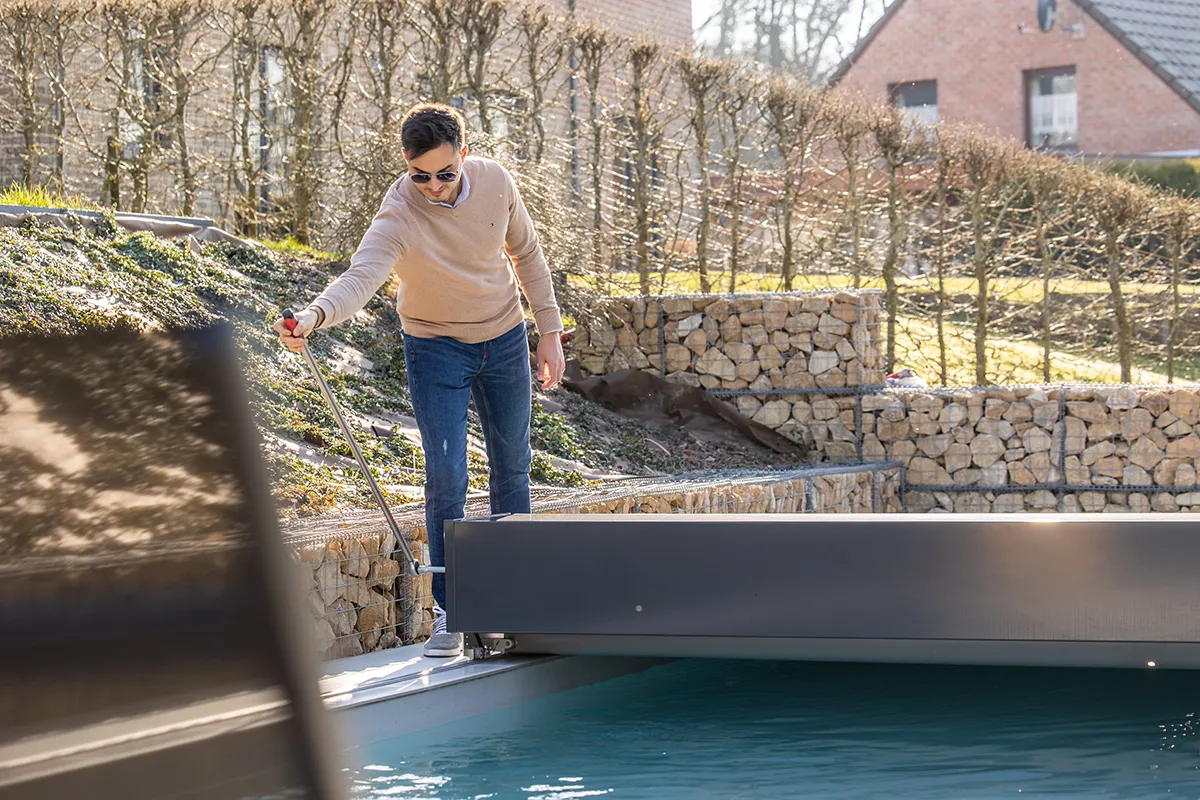 Un homme en pull beige manipule un outil près d'une piscine extérieure entourée de haies et de murs en pierre.