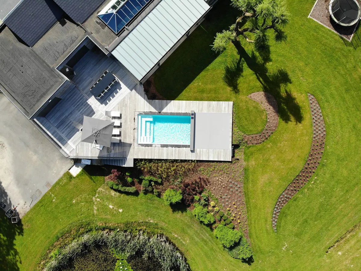 Vue aérienne d'une terrasse en bois avec piscine rectangulaire, parasol gris et jardin verdoyant autour.