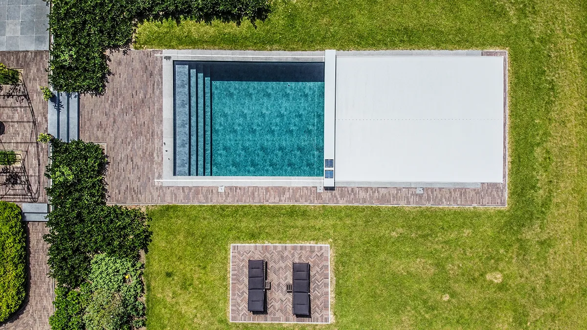 Une piscine rectangulaire avec couverture blanche partielle est entourée d'un pavage en pierre et de pelouse verte.