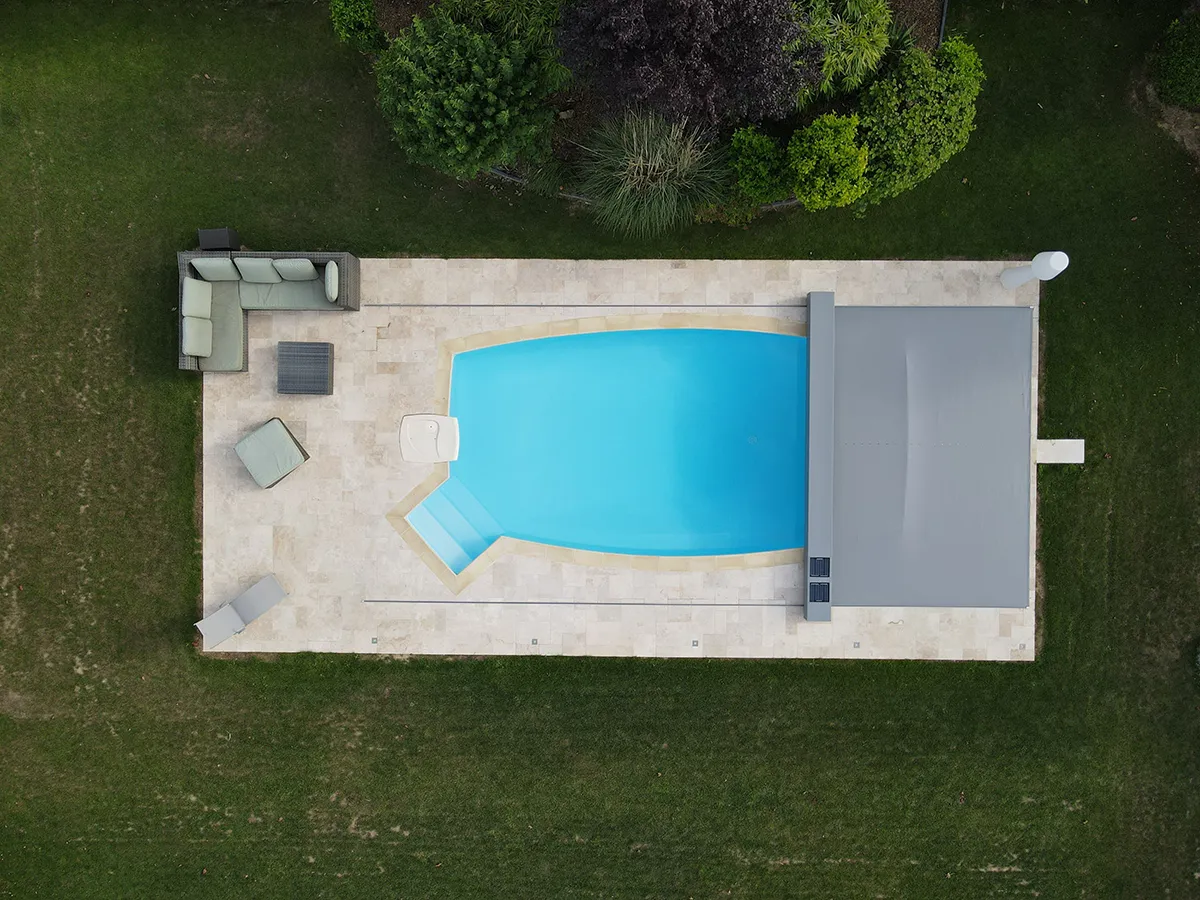 Vue aérienne d'une piscine bleue entourée de dalles blanches, avec des meubles de jardin gris sur la terrasse et pelouse verte autour.