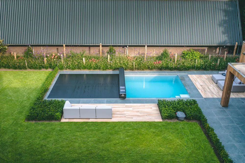 Piscine rectangulaire entourée de haies vertes, avec une terrasse en bois et des canapés gris, située près d'un bâtiment métallique.