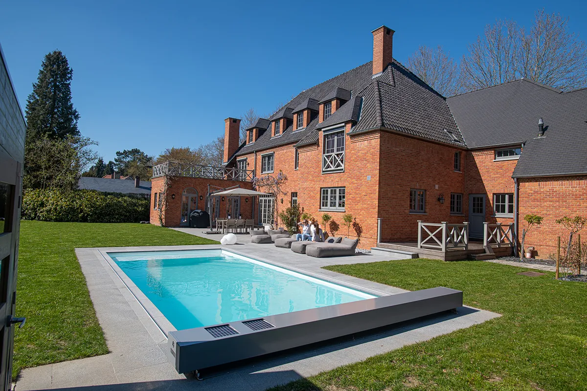 Grande maison en briques rouges avec piscine rectangulaire et mobilier de jardin gris, entourée d'espaces verts et d'arbres.