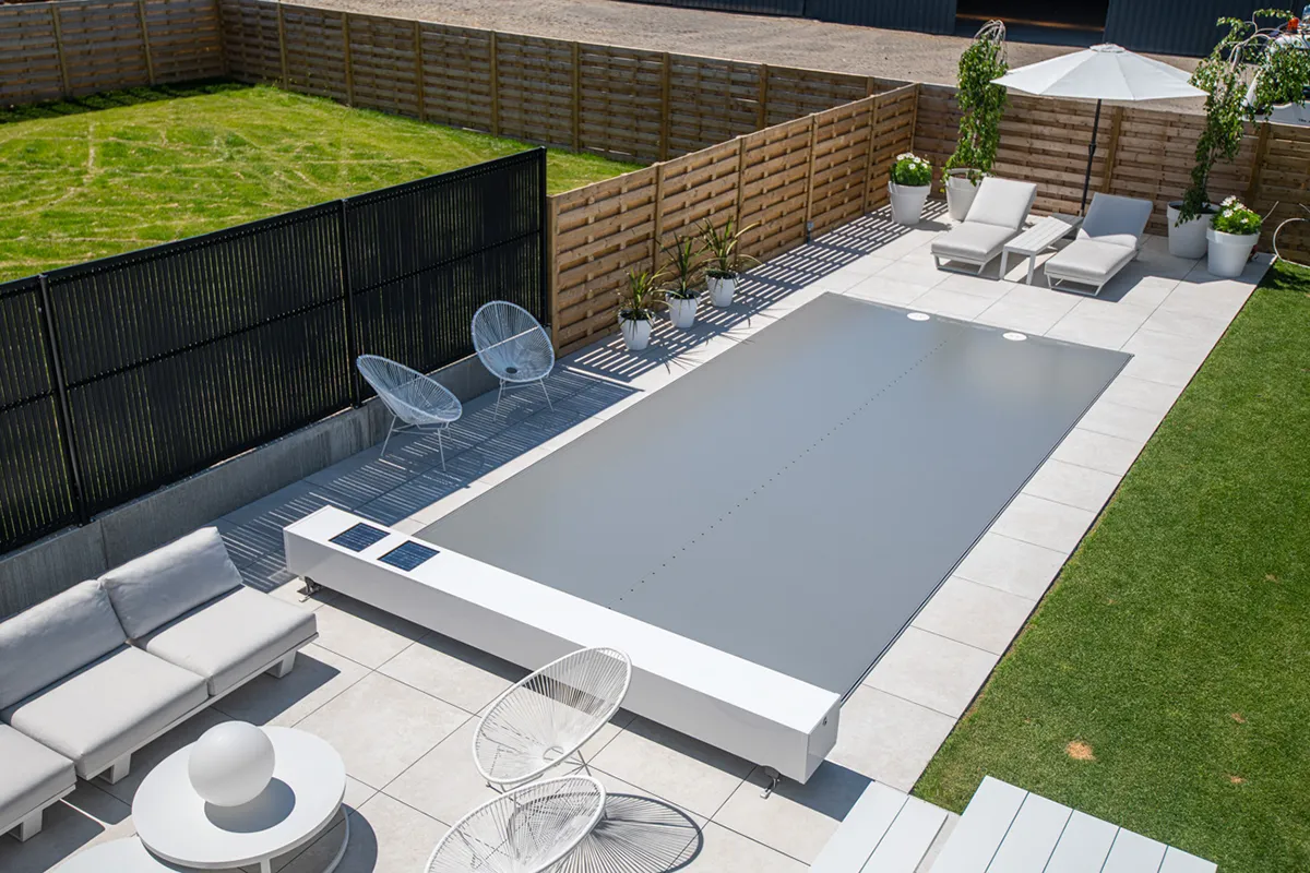 Une piscine couverte d'une bâche grise est entourée de chaises blanches, de plantes en pots et d'un parasol blanc sur une terrasse.