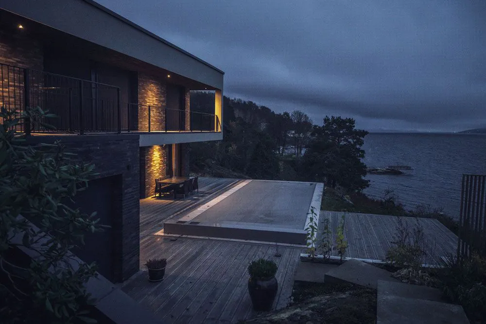 Maison moderne avec terrasse en bois éclairée, piscine couverte, vue sur un lac et ciel nuageux en arrière-plan.