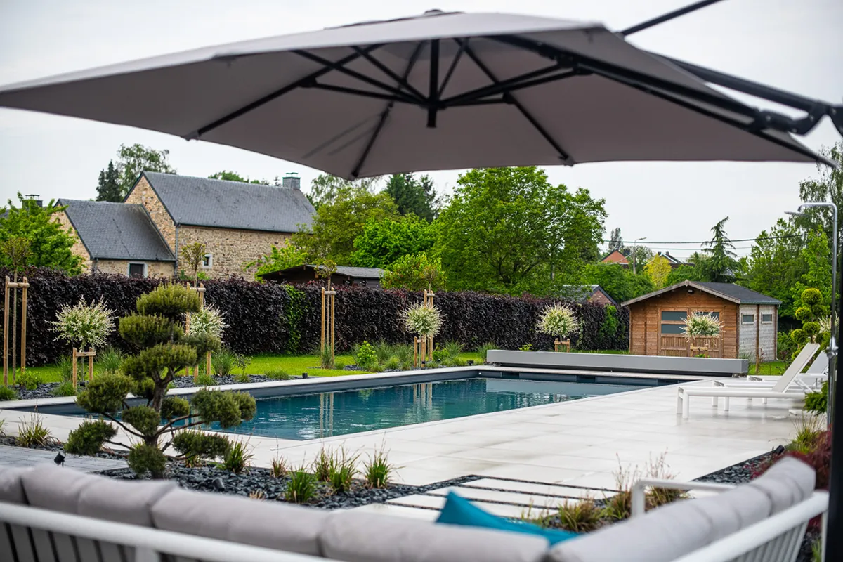 Une piscine entourée de dalles blanches, avec des chaises longues, un parasol gris, des arbustes et un cabanon en bois en arrière-plan.
