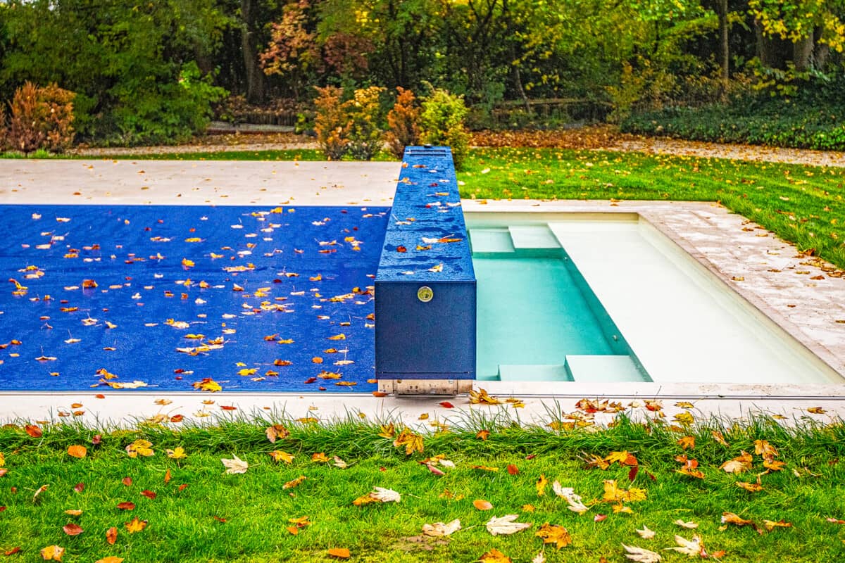 BLUE-COVER-(1) Couverture de piscine bleue avec feuilles mortes, entourée de pelouse verte et arbres en arrière-plan.