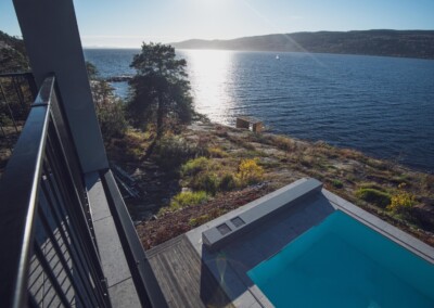 Vue d'un balcon surplombant une piscine bleue bordée de dalles grises, avec un paysage côtier et un arbre en arrière-plan.